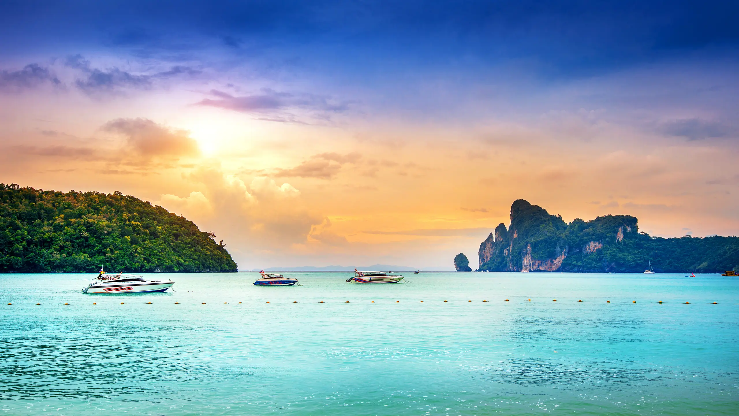 Speedboats in a turquoise bay framed by jungle-covered limestone islands at sunset, Thailand