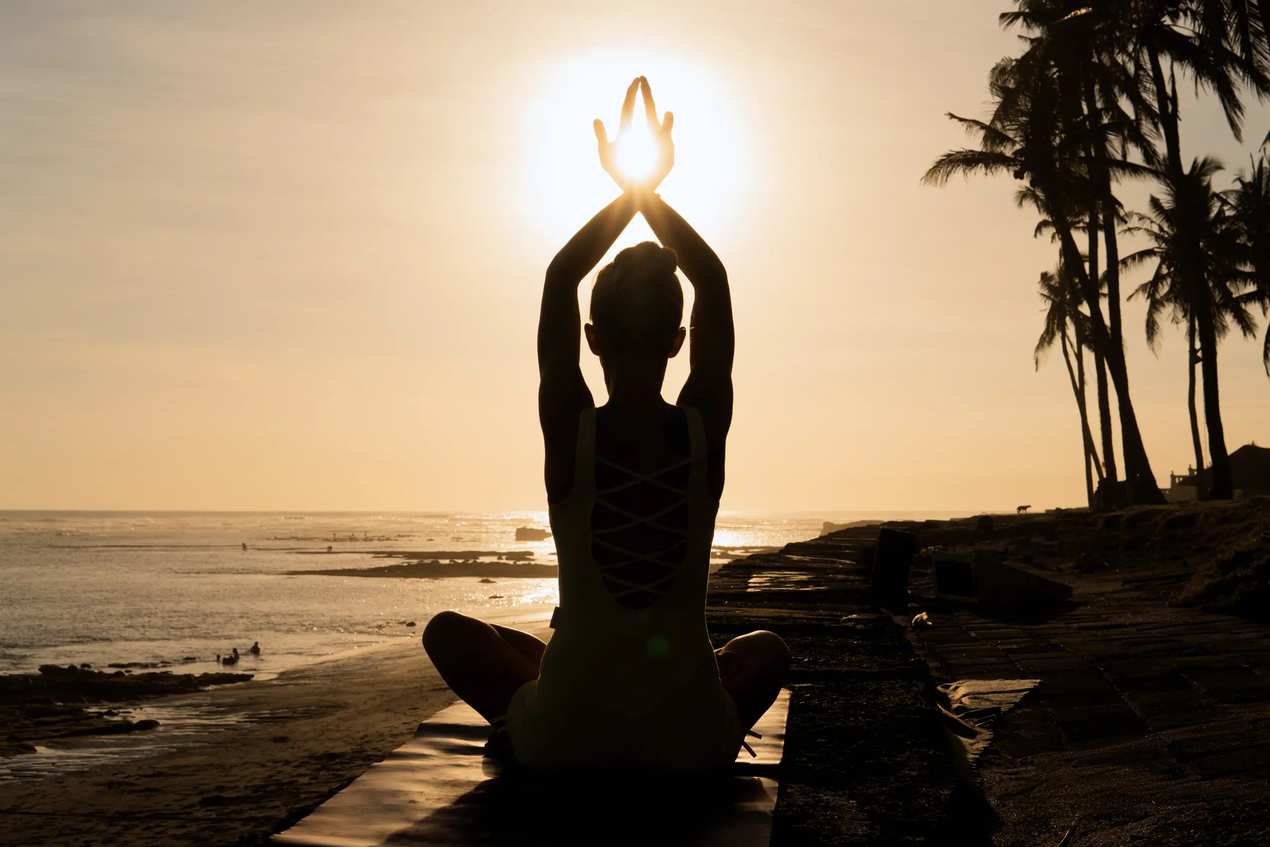 Silhouette of a woman meditating cross-legged on a seaside path at sunset, hands joined above her head